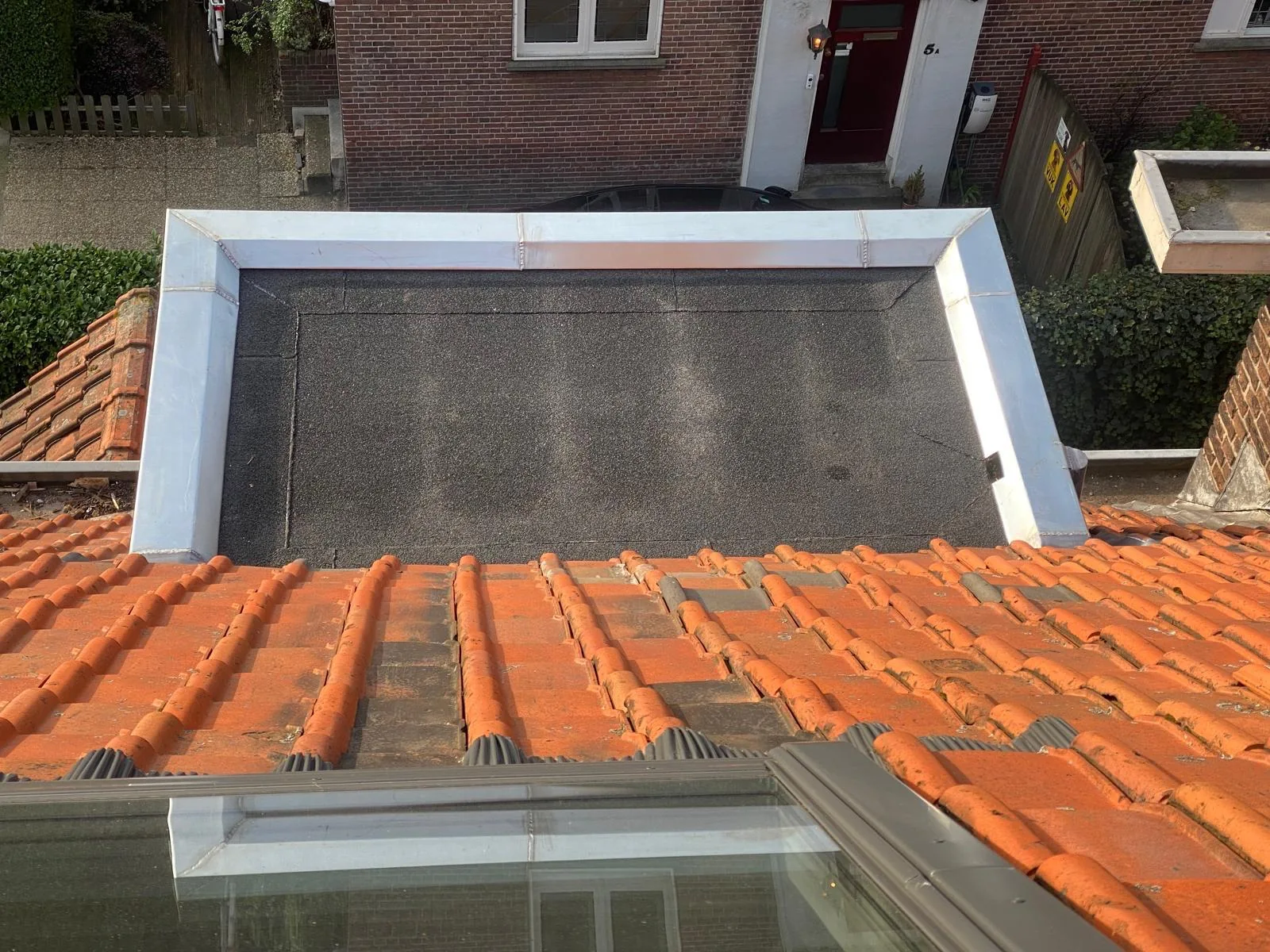 Klein plat dak met metalen randafwerking naast een rood pannendak.